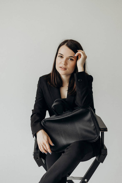 A girl in a black suit with a bag on a white background sits on a chair - Fotografie, Obrázek