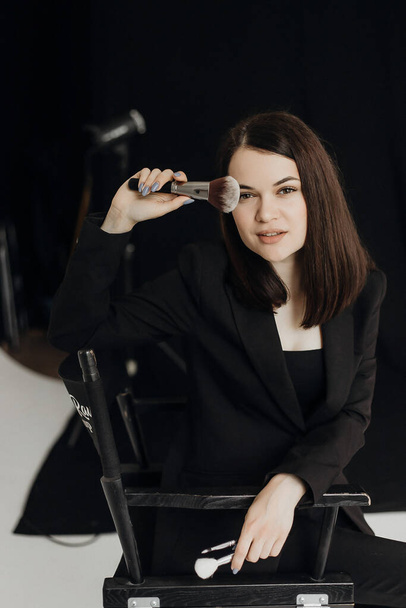 A girl in a black suit with a brush in her hands on a black background sits on a chair - Foto, immagini