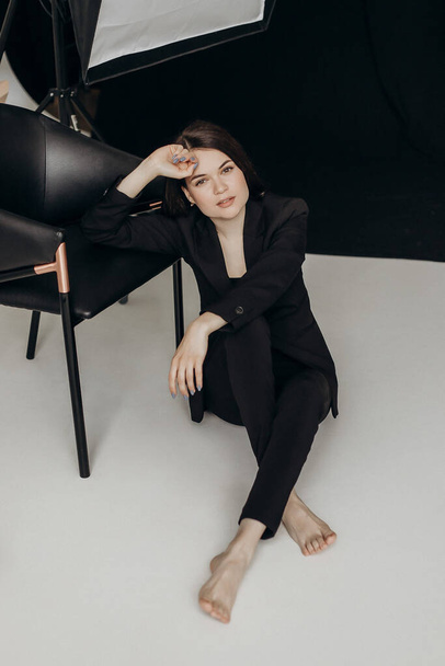 A girl in a black suit on a white background sits on the floor near a chair - Zdjęcie, obraz