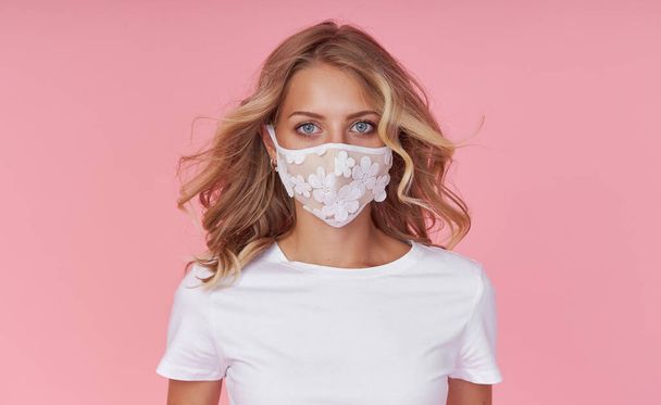 A young attractive woman in a white casual t-shirt wears a protective face mask. Protection against flu and cold diseases during a pandemic. Pink isolated background - Photo, Image