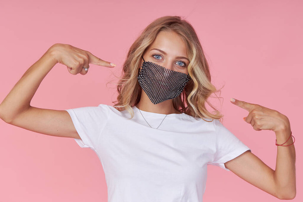 A young attractive woman in a white casual t-shirt wears  a stylish fashionable protective face mask. Protection against flu and cold diseases during a pandemic. Pink isolated background - Foto, Imagem