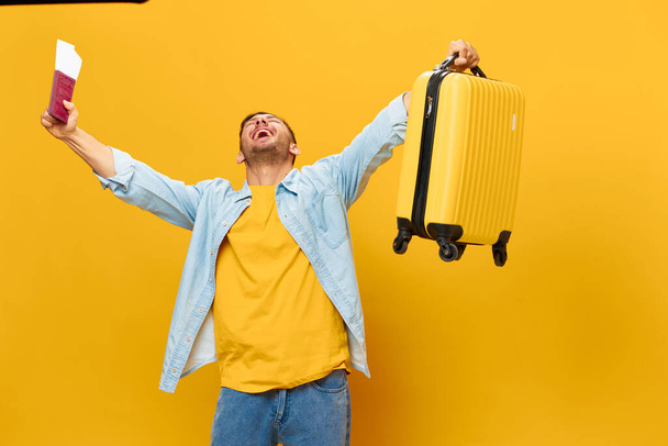 Aşırı mutlu, bronzlaşmış, mavi gömlekli yakışıklı adam, sarı stüdyo arka planında izole bir şekilde duran pasaport uçak biletleriyle ve bavulla el kaldırsın. Uzayın afiş modelini kopyala. Seyahat konsepti - Fotoğraf, Görsel