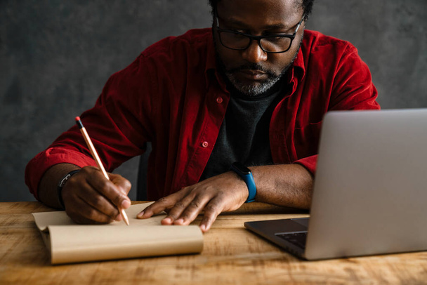 Black man in eyeglasses writing down notes while working with laptop indoors - Photo, Image