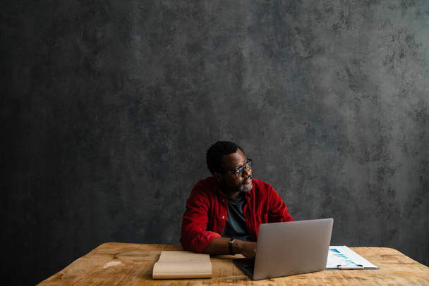 Black man in eyeglasses working with laptop while sitting at desk indoors - Foto, immagini