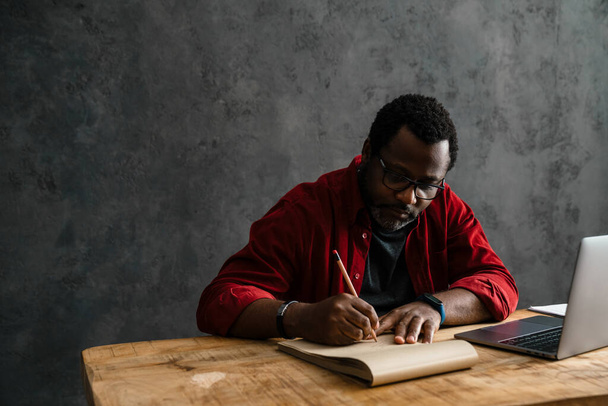 Black man in eyeglasses writing down notes while working with laptop indoors - Фото, зображення