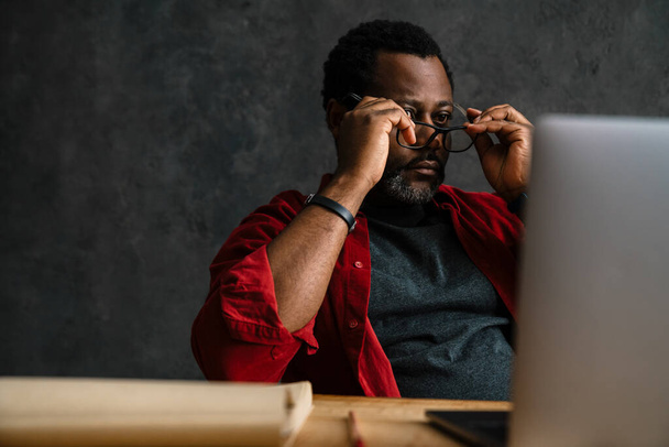 Black man in eyeglasses working with laptop while sitting at desk indoors - 写真・画像