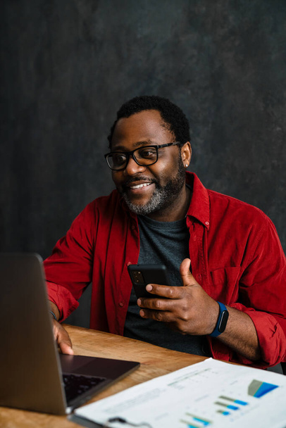 Black man in eyeglasses using cellphone while working with laptop indoors - Foto, imagen
