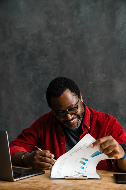 Black man in eyeglasses smiling while working with laptop and papers indoors - Foto, Imagen