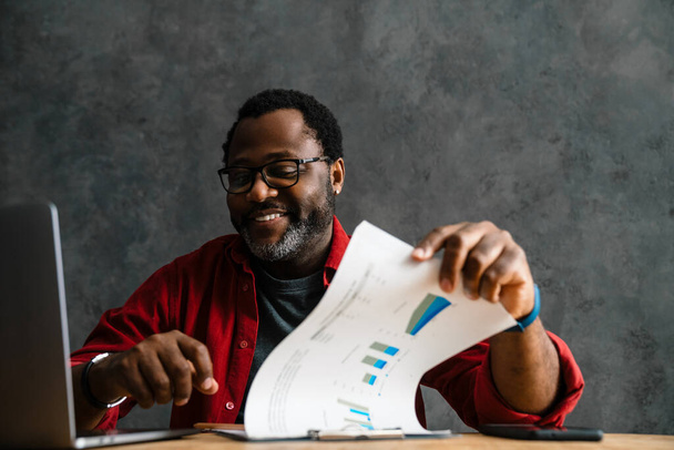 Black man in eyeglasses smiling while working with laptop and papers indoors - Fotó, kép