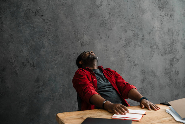 Black tired man in eyeglasses working with laptop while sitting at desk indoors - Fotografie, Obrázek
