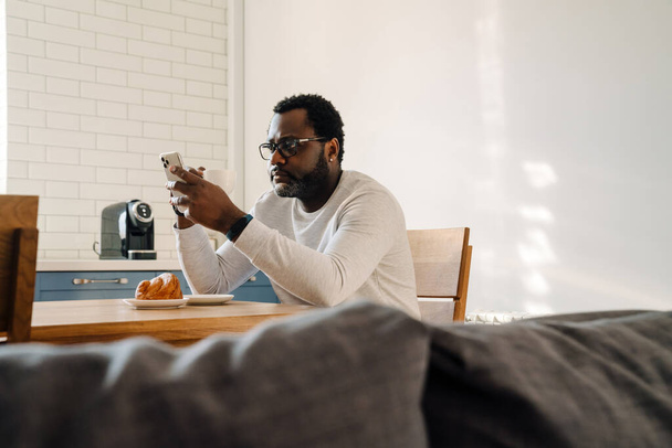 Black man wearing eyeglasses using mobile phone while having breakfast at home - Fotoğraf, Görsel