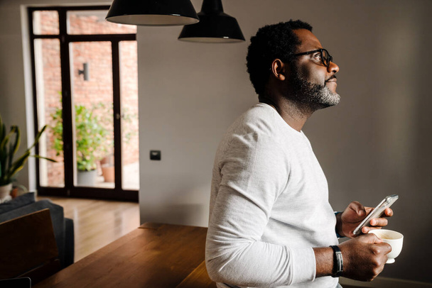 Black man wearing eyeglasses using mobile phone while drinking coffee at home - Foto, Bild