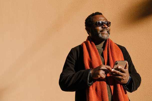 Black bearded man in sunglasses using cellphone while standing by wall outdoors - Foto, Bild