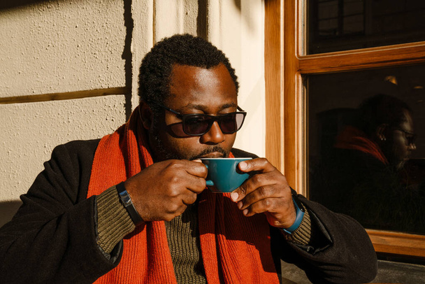 Black bearded man wearing sunglasses drinking coffee while sitting in street cafe outdoors - Фото, изображение