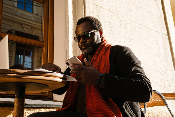 Чорний бородатий чоловік використовує мобільний телефон під час роботи в кафе на відкритому повітрі
 - Фото, зображення