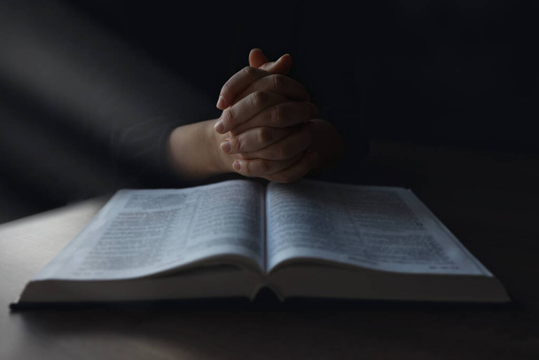 Las manos de la mujer en la Biblia. está leyendo y rezando sobre la Biblia en un espacio oscuro sobre una mesa de madera
. - Foto, Imagen