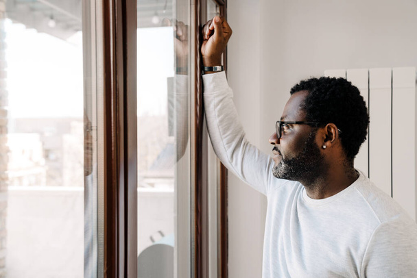 Black bearded man wearing eyeglasses looking in window at home - Foto, imagen