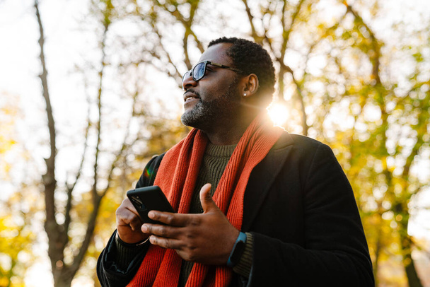 Black bearded man in sunglasses using cellphone while strolling at park - Photo, image