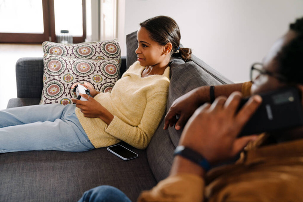 Black girl playing video game while her father talking on cellphone at home - Foto, afbeelding