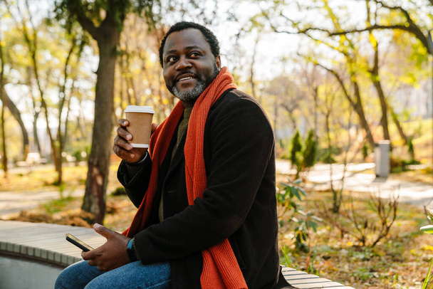 Black man using cellphone and drinking coffee while sitting on bench in park - Foto, immagini