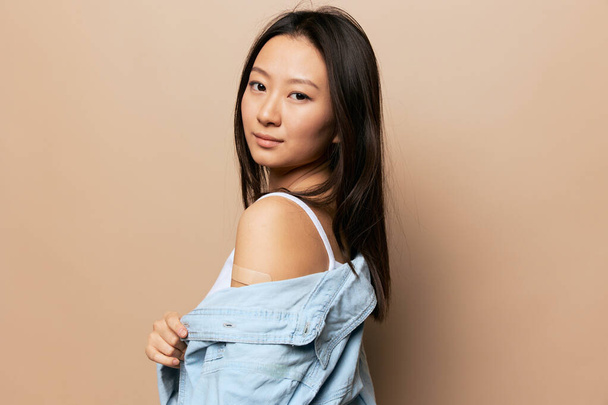 Portrait of happy tanned adorable young Asian lady after vaccination hold shirt sleeve posing isolated on beige pastel background. People Emotions. CoVID Vaccination concept. Copy Space Offer Banner. - Foto, Imagen