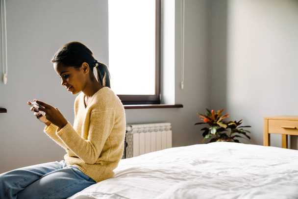 Black brunette girl using mobile phone while sitting on bed at home - Foto, Imagen