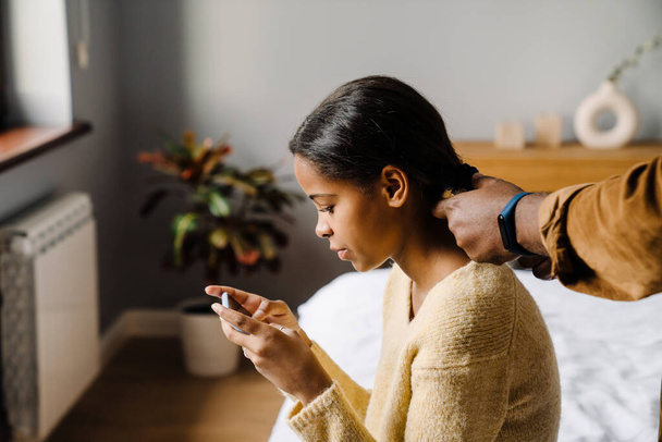 Black girl using cellphone while father doing her hairstyle at home - Valokuva, kuva