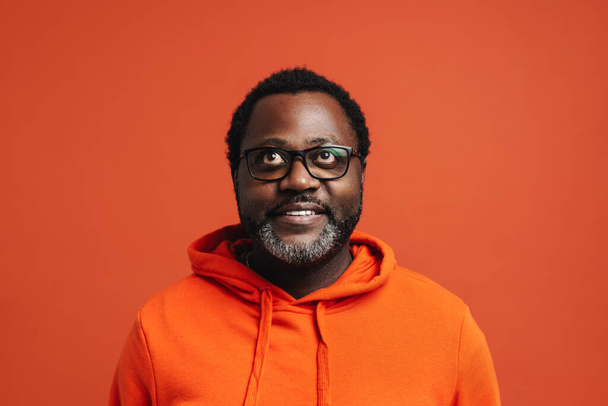 Black bearded man in eyeglasses smiling and looking upward isolated over orange background - Foto, immagini