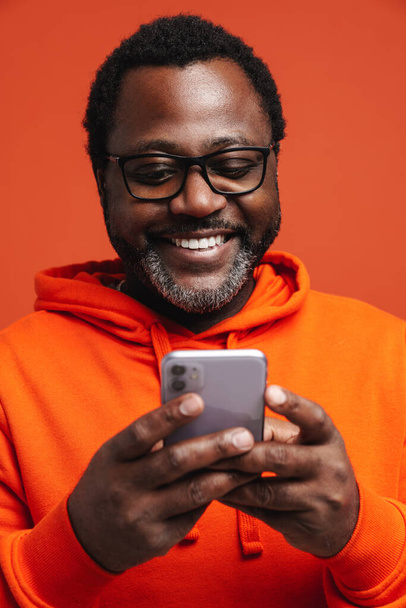 Black bearded man in eyeglasses smiling and using cellphone isolated over orange background - Foto, afbeelding