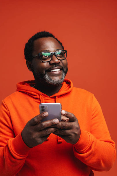 Black bearded man in eyeglasses smiling and using cellphone isolated over orange background - Fotó, kép