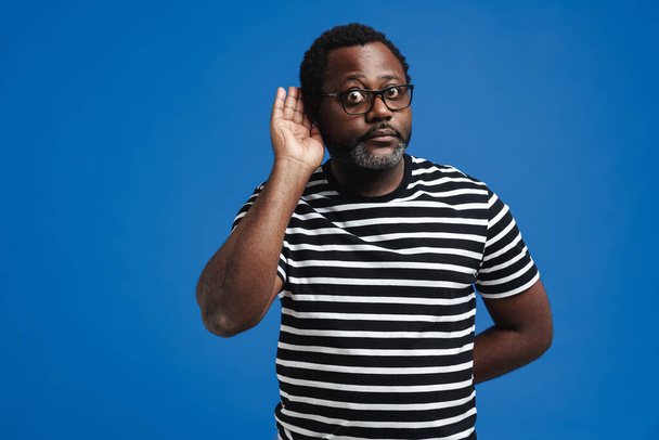 Black bearded man expressing surprise while listening isolated over blue background - Foto, afbeelding