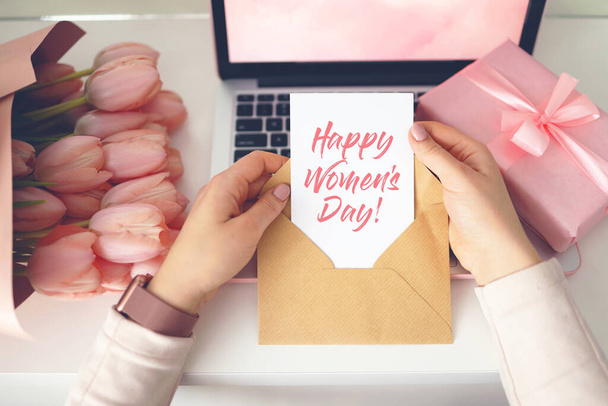 Woman's hands holding a letter in craft envelope. Pink background, women's day concept. Tulips flower and pink gift box in background. Womens home desk. - Photo, image