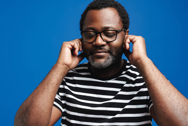 Black man in eyeglasses smiling while listening music with earphones isolated over blue background - Fotó, kép