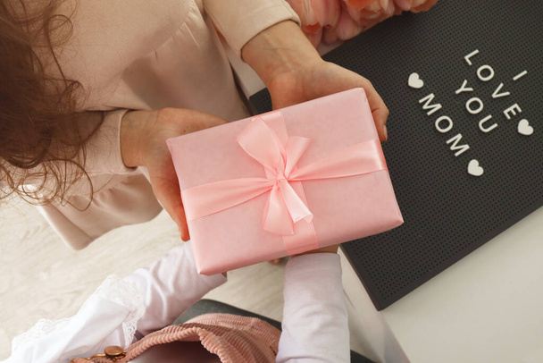 Child's hands hold beautiful pink gift box with ribbon. Pink tulip flower and sign i love you mom in background. Top view, close-up. Preparing for the holidays. - Фото, изображение