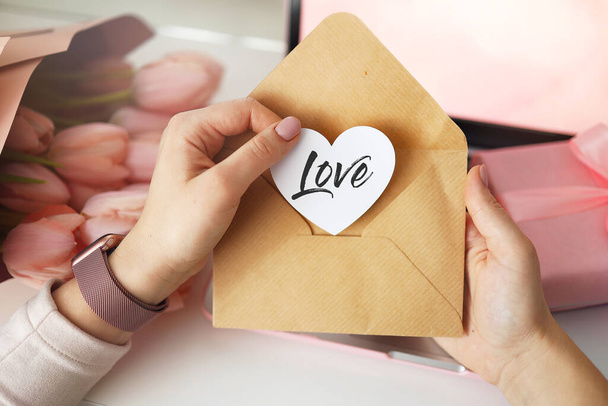 Woman's hands holding a letter in craft envelope. Pink background, valentine's day concept. Tulips flower and pink gift box in background. Womens home desk. - Foto, Bild
