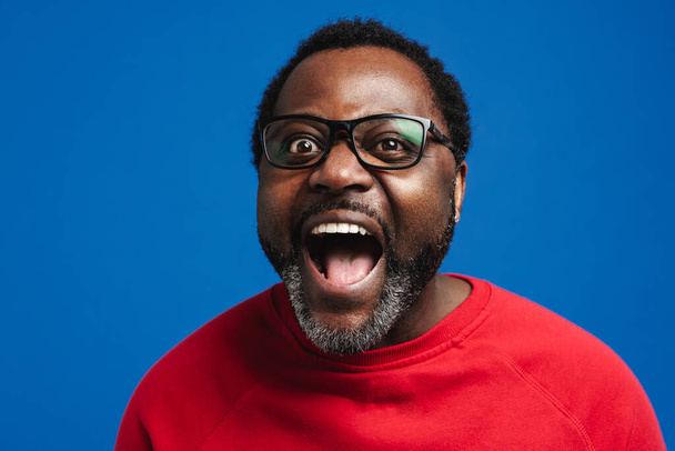 Black bearded man in eyeglasses expressing surprise at camera isolated over blue background - Фото, изображение
