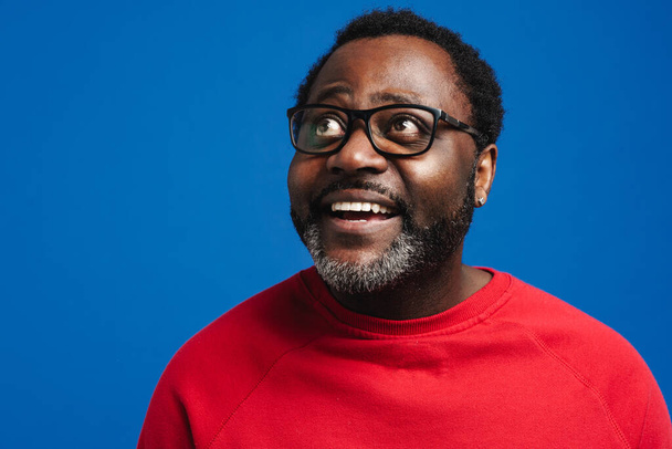 Black bearded man in eyeglasses expressing surprise at camera isolated over blue background - Фото, зображення