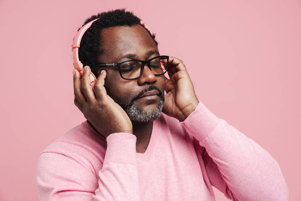 Black bearded man in eyeglasses listening music with headphones isolated over pink background - Foto, afbeelding