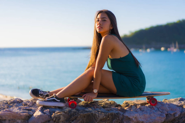 A young brunette latin woman sitting on a skateboard enjoying summer by the sea - Foto, Imagen