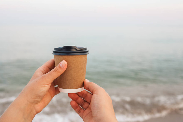 Coffee at the sea. Young woman holds a disposable cup of coffee on on a sunny morning at the beach. - Φωτογραφία, εικόνα