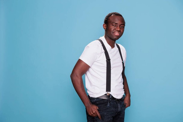 Confident man posing for camera while standing on blue background. Smiling heartily handsome young adult person with fashion style wearing white tshirt and suspenders. Studio shot - Zdjęcie, obraz