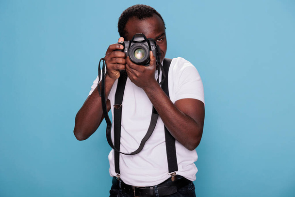 African american photography enthusiast taking photo while standing on blue background. Confident professional photographer having DSLR camera taking picture. Studio shot. - Fotó, kép