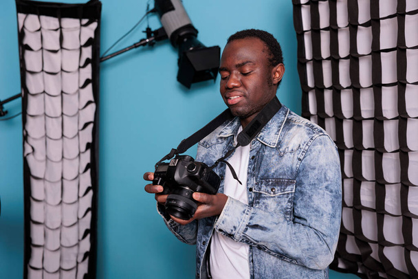 Happy photography enthusiast with DSLR device standing on blue background. Smiling photographer checking camera gallery while standing in production studio equipped with softboxes and spotlights. - Foto, afbeelding