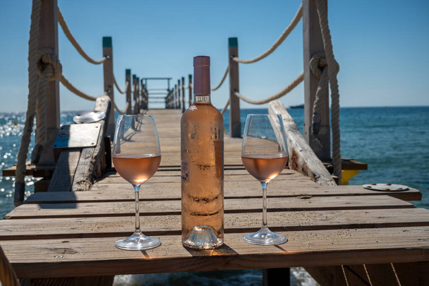 Glasses and bottle of cold rose wine from Provence served outdoor on wooden yacht pier with view on blue water and white sandy beach Plage de Pampelonne near Saint-Tropez, summer vacation in France - Fotoğraf, Görsel