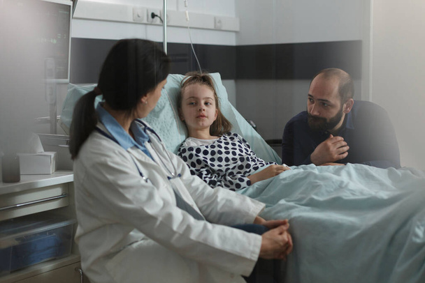 Pediatrician examining sick girl disease symptoms while resting in patient bed. Doctor expert discussing with girl about her health condition while caring father sitting beside ill daughter. - Fotografie, Obrázek