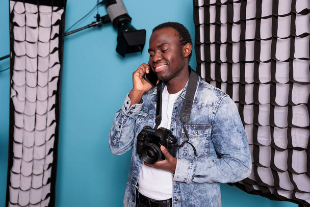 Young producer having a phone call while working on production studio equipped with professional softboxes and spotlights. Photographer talking on smartphone while standing on blue background. - Fotó, kép
