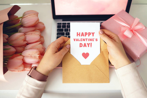 Woman's hands holding a letter in craft envelope. Pink background, valentine's day concept. Tulips flower and pink gift box in background. Womens home desk. - Foto, Imagen