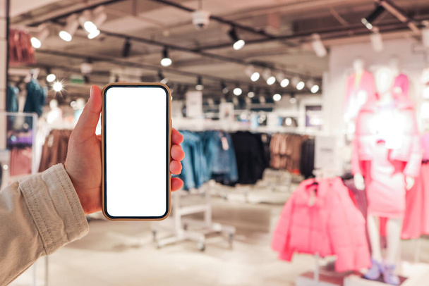 A girl holds a phone in her hand with a blank isolated screen on the background of a blurred shopping center. - Foto, Imagem