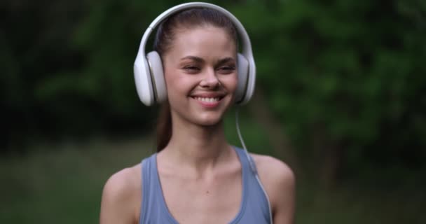 Femme portant de grands écouteurs blancs écoute sa musique préférée tout en s'entraînant dans la nature et sourit avec bonheur et style de vie. Images 4k de haute qualité - Séquence, vidéo