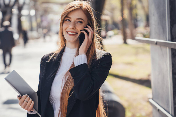 Smiling young caucasian red-haired businesswoman talking on mobile phone, outdoors in the park. Phone communication. Business call. - Foto, afbeelding